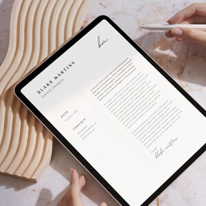 May include: A digital tablet displaying a resume for Blake Martins, Interior Designer. The tablet is held by a person's hands, with a stylus nearby. The background includes a wooden decorative piece and a marble surface.