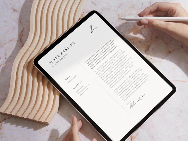 May include: A digital tablet displaying a resume for Blake Martins, Interior Designer. The tablet is held by a person's hands, with a stylus nearby. The background includes a wooden decorative piece and a marble surface.