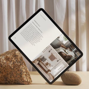 May include: A digital tablet displaying a portfolio of interior design work. The tablet is propped up on a rock and shows a photo of a living room with a sofa, a bench, and a rug. The text on the tablet reads "Portfolio 2:  Coastal Retreat Renovation".