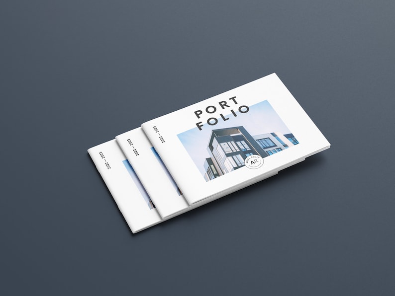 Puede incluir: Una pila de libros de portafolio blancos con la palabra "PORTFOLIO" en letras negras en negrita. La portada presenta una imagen de un edificio moderno. Los libros est&aacute;n etiquetados "2022-2023" en el lomo y est&aacute;n colocados sobre una superficie gris oscuro.