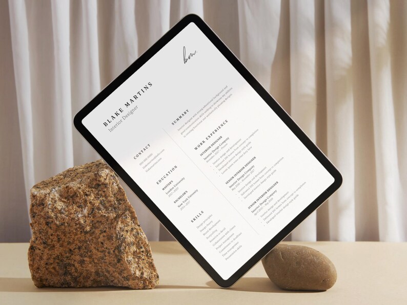 May include: A digital tablet displaying a resume for "Blake Martins, Interior Designer." The resume includes sections for contact, education, skills, and work experience. The tablet is propped up by a rock and a small stone against a neutral background.