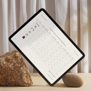 Pode incluir: Um tablet branco com moldura preta est&aacute; apoiado em uma rocha. A tela exibe uma programa&ccedil;&atilde;o de m&oacute;veis e decora&ccedil;&atilde;o com uma lista de itens, quantidades e pre&ccedil;os. O texto "Furniture & Decor Schedule" &eacute; vis&iacute;vel na parte superior da tela.