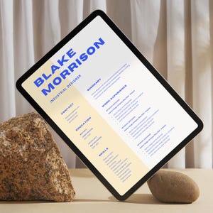 May include: A digital tablet displaying a CV for Blake Morrison, an Industrial Designer. The CV includes sections for contact information, education, work experience, skills, and a summary. The tablet is propped up on two rocks.