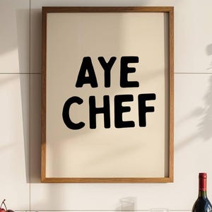 May include: A framed wall print with a cream background and the black text AYE CHEF in a bold, rounded font. The frame is made of medium-toned wood. The print is displayed above a kitchen counter with a wooden bowl of fruit and a wine bottle.