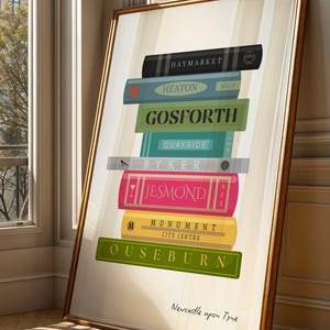 Może przedstawiać: Oprawiony wydruk przedstawiający stos kolorowych książek z nazwami miejsc, takimi jak Gosforth i Jesmond. Wydruk ma złotą ramę i opiera się o ścianę. Tekst "Newcastle upon Tyne" znajduje się na dole.