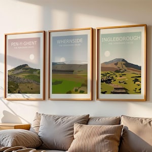 May include: Three framed landscape prints with the names "PEN-Y-GHENT", "WHERNSIDE", and "INGLEBOROUGH" are displayed above a gray sectional sofa. The prints feature mountain scenes with a minimalist design. A wooden cabinet and decorative items are also visible.