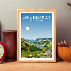 Op de afbeelding: Een ingelijste print van een landschapsscène met een meer en bergen. De print heeft de tekst "LAKE DISTRICT NATIONAL PARK" bovenaan.