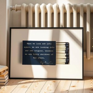 May include: A framed rectangular print featuring a dark blue matchbox graphic with a row of matches on the right side. The text reads: When we look out into space, we are looking into our own origins, because we are truly children of the stars.