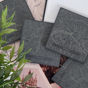 May include: Four black square coasters with engraved city maps. The coasters feature the names "Rome", "London", "Paris", and "Amsterdam" in white lettering.