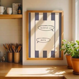 May include: A framed print with a light wood frame and a cream background with vertical navy blue stripes. The print has two speech bubbles with the words "kettle's on" and "hero". The print is displayed in a kitchen setting.