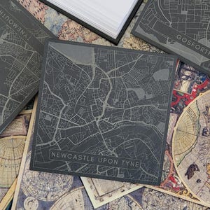 May include: A black slate coaster featuring a white outline map of Newcastle upon Tyne, England. The coaster is displayed on a background of vintage maps.