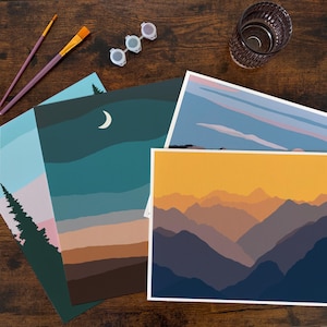 Op de afbeelding: Een verzameling landschapskunstprints met schildersbenodigdheden. De prints tonen berg- en luchtscènes in tinten blauw, oranje en bruin. Penselen, verfpotten en een glas water zijn ook zichtbaar.