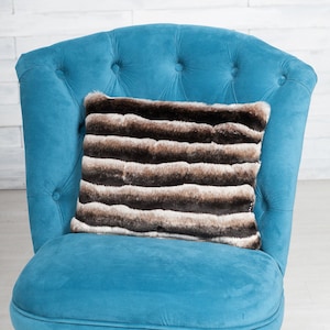 Op de afbeelding: Een turquoise fluwelen fauteuil met een gecapitonneerde rugleuning en donkere houten poten. Een rechthoekig kussen met horizontale strepen van bruin, beige en wit imitatiebont ligt op de zitting.
