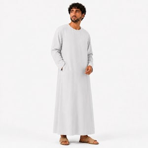 May include: A light gray, floor-length thobe featuring long sleeves, a round neckline, and a straight-cut silhouette. The garment is constructed with a simple, clean design, including side pockets, and is worn with tan open-toe sandals.