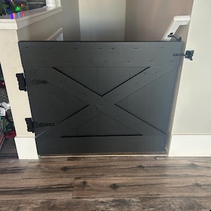 May include: Black wooden gate with an X-shaped design, attached to a white wall with black hinges and a white latch.