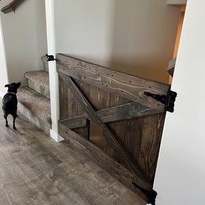 May include: A dark brown wooden pet gate with a diagonal X design and a small pet door. The gate is attached to a white wall with black hinges.