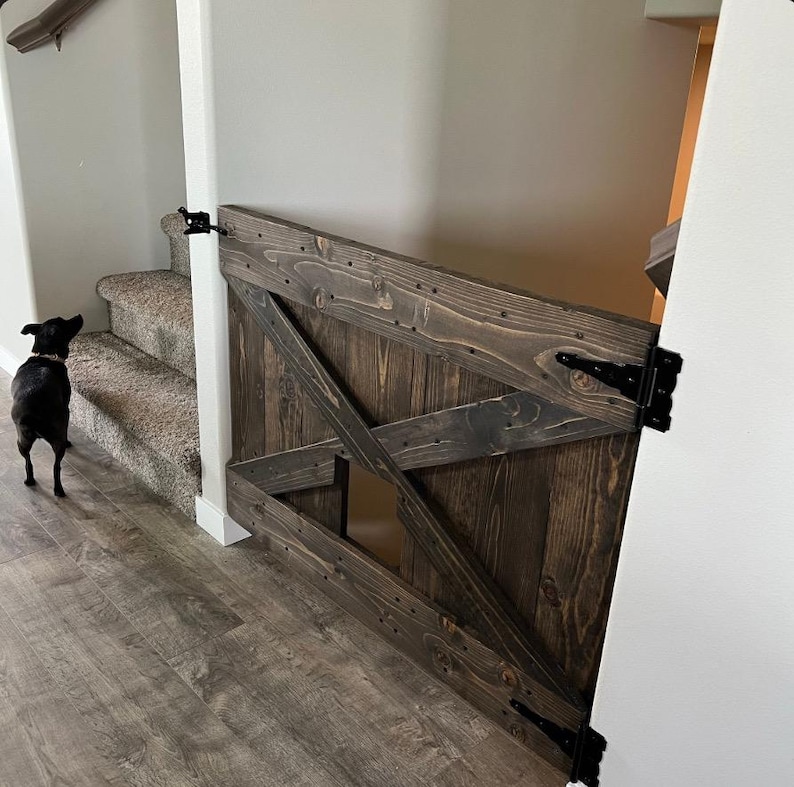 May include: A dark brown wooden pet gate with a diagonal X design and a small pet door. The gate is attached to a white wall with black hinges.