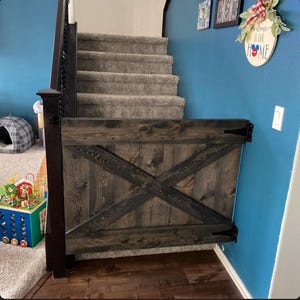 May include: A dark wood baby gate with a diagonal cross design, mounted to a wall and stairs. The gate is closed and has black metal hinges. The wall is painted a teal blue color.