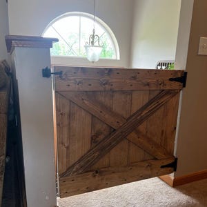 May include: A wooden gate with a rustic, distressed finish and a diagonal X-shaped design. The gate is hinged on the right side and has black metal hinges. The gate is closed and appears to be a pet gate.