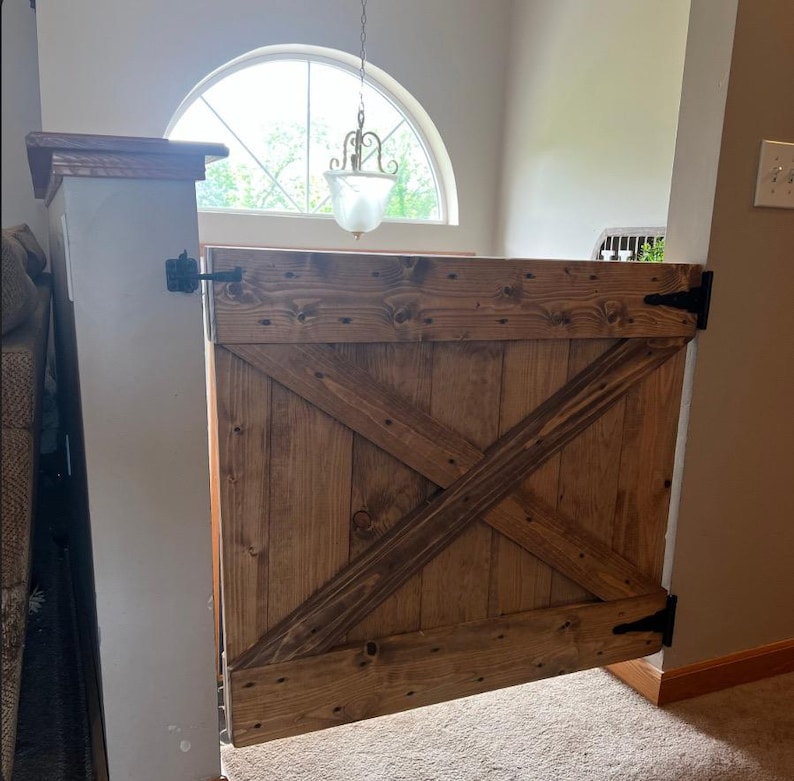 May include: A wooden gate with a rustic, distressed finish and a diagonal X-shaped design. The gate is hinged on the right side and has black metal hinges. The gate is closed and appears to be a pet gate.