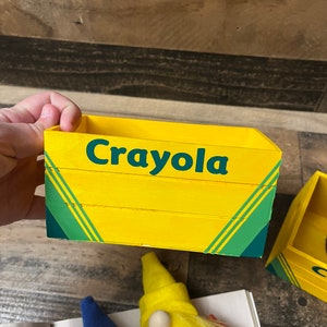 May include: A yellow wooden box with the word "Crayola" printed on the front. The box has green stripes and is sitting on a wooden surface.