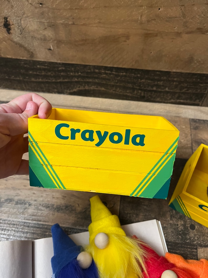 May include: A yellow wooden box with the word "Crayola" printed on the front. The box has green stripes and is sitting on a wooden surface.