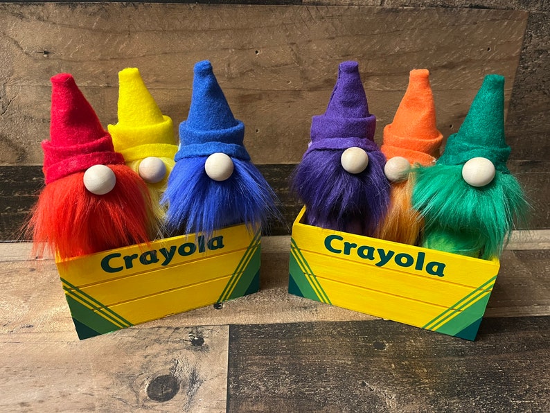 May include: Five gnome figurines in a yellow Crayola box. The gnomes are dressed in different colors of felt, resembling crayons. The gnomes are arranged in a row, with the red gnome on the left and the green gnome on the right.