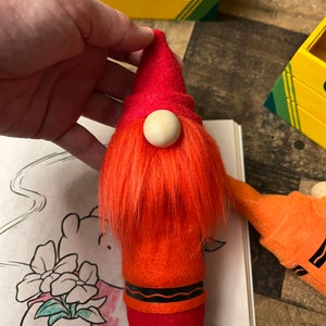 May include: A red felt gnome with a white wooden nose and orange fuzzy hair. The gnome is shaped like a crayon and has a black wavy line around the bottom.