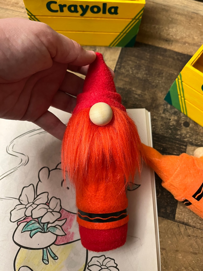May include: A red felt gnome with a white wooden nose and orange fuzzy hair. The gnome is shaped like a crayon and has a black wavy line around the bottom.
