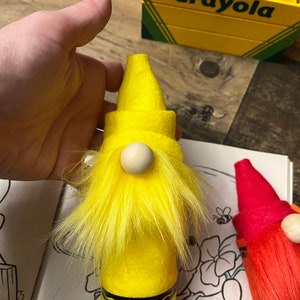 May include: A yellow crayon gnome with a white wooden nose and yellow fur. The gnome is sitting on a coloring book page with a flower and a bee.
