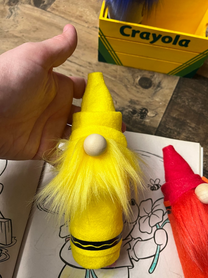 May include: A yellow crayon gnome with a white wooden nose and yellow fur. The gnome is sitting on a coloring book page with a flower and a bee.