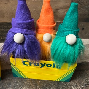 May include: Three gnomes with purple, orange, and green felt hats and fuzzy white beards sitting in a yellow wooden box with the word "Crayola" printed on the front.