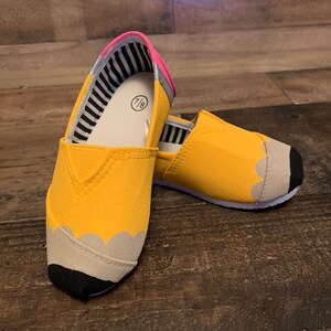 May include: A pair of yellow canvas slip-on shoes with a pencil design. The shoes have a black eraser tip, a gray pencil lead, and a pink and gray striped lining. The size 7/8 is visible on the inside of the shoe.