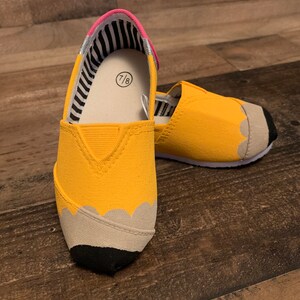 May include: A pair of yellow canvas slip-on shoes with a pencil design. The shoes have a black and white striped lining and a pink trim. The size 7/8 is visible on the inside of the shoe.