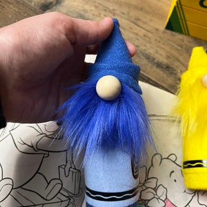 May include: Blue crayon gnome with a blue felt hat and a wooden nose. The gnome is standing on a white surface with a coloring book in the background.