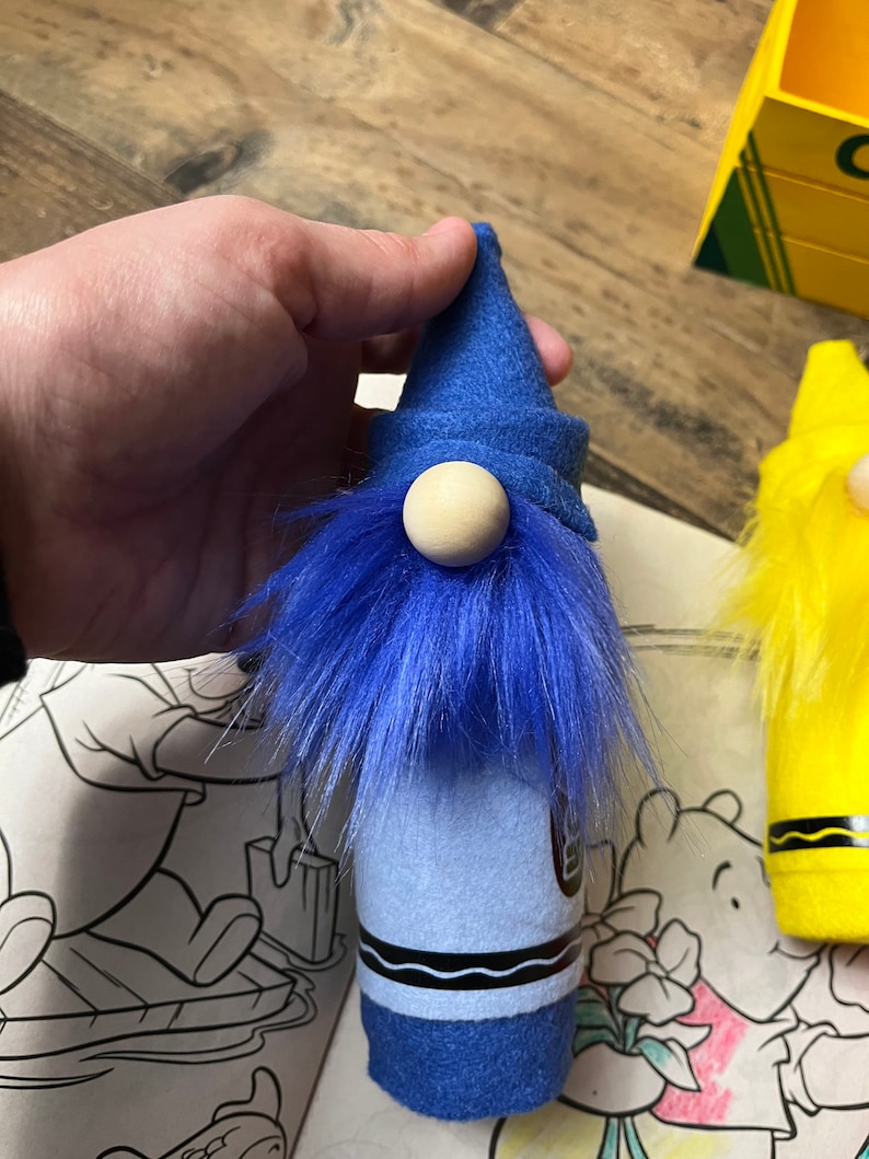 May include: Blue crayon gnome with a blue felt hat and a wooden nose. The gnome is standing on a white surface with a coloring book in the background.