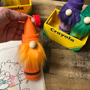 May include: Orange, purple, and green crayon gnomes with white beards and hats. The gnomes are sitting in a yellow wooden box with the word "Crayola" printed on the front.