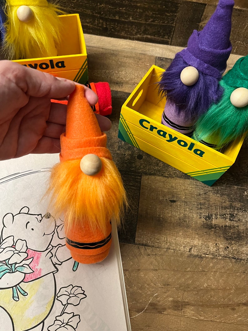 May include: Orange, purple, and green crayon gnomes with white beards and hats. The gnomes are sitting in a yellow wooden box with the word "Crayola" printed on the front.