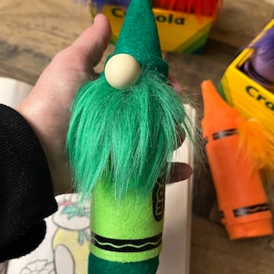 May include: A green crayon gnome with a white pom-pom nose and green fuzzy beard. The gnome is standing on a book with a drawing of a cartoon character.
