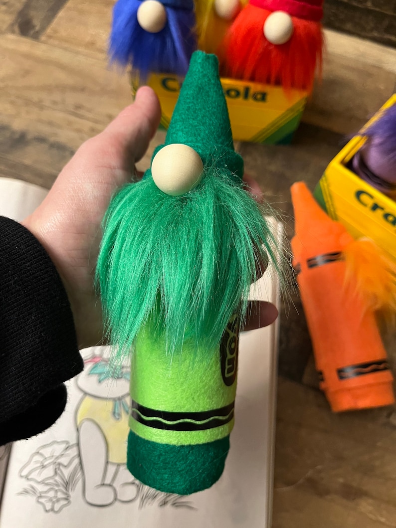 May include: A green crayon gnome with a white pom-pom nose and green fuzzy beard. The gnome is standing on a book with a drawing of a cartoon character.