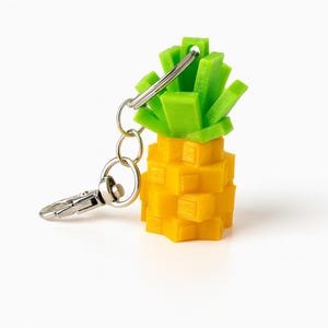 May include: A yellow and green pineapple-shaped keychain. The keychain is made of a 3D-printed material and features a silver metal clasp and key ring. The pineapple has a textured surface and a bright, cheerful appearance.