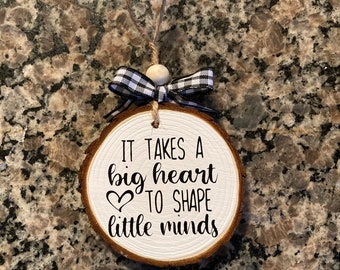 May include: A round wooden ornament with the text "IT TAKES A big heart TO SHAPE little minds" in black script. The ornament has a white painted surface, a black and white checkered bow, and a twine loop for hanging.
