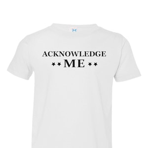 May include: White toddler t-shirt with black text that reads "ACKNOWLEDGE ME" with three stars on each side of the word "ME".