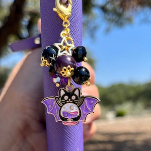 May include: A gold charm with a black cat wearing a purple bat costume holding a potion bottle with a crescent moon and stars. The charm is attached to a purple lanyard with black and purple beads and gold stars.
