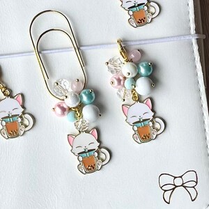 May include: Four gold-tone cat charms with a boba tea design. Each charm is attached to a different type of bookmark, including a clip, a chain, and a beaded string.