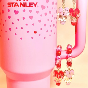 May include: A pink Stanley travel mug with a heart pattern and the Stanley logo. The mug has a handle with two charms attached. The charms are gold-colored and feature a heart with the word "LOVE" on it. The charms are attached to a chain of small pink and red beads.