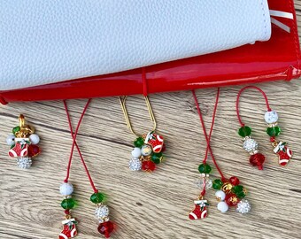 May include: A set of five Christmas-themed bookmarks with red, green, and white beads, charms, and a stocking design. The bookmarks are attached to a red leather journal.