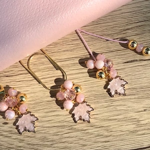 May include: Three pink and gold leaf charms with pink beads and a gold clip. The charms are on a wooden surface.