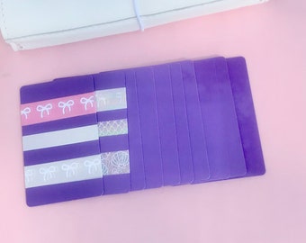 May include: A stack of purple cardstock paper with decorative white and pink washi tape on the top card. The cardstock is blank and ready for crafting.