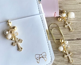 May include: Two gold cross charms with white pearl and crystal accents. The charms are attached to a white leather notebook and a gold paper clip.